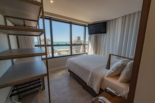 Quarto de hotel com cama, escrivaninha, televisão e vista para a praia e oceano.