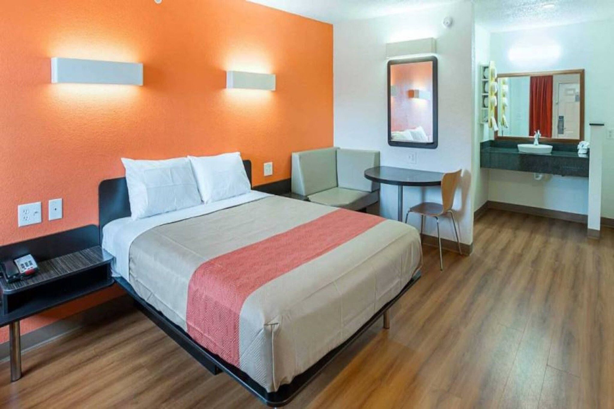 A hotel room with a bed, a desk, a chair, a small table, and a bathroom with a mirror and sink.