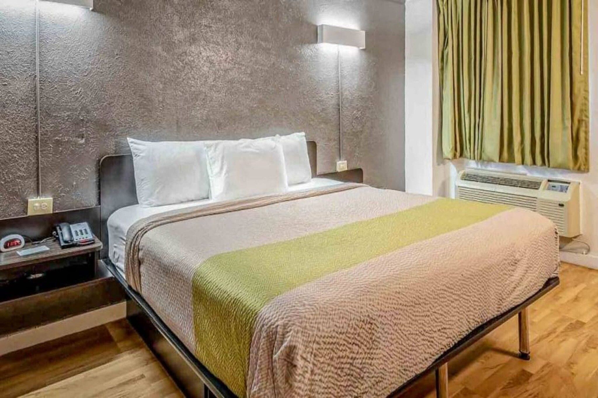 A hotel room with a bed, bedside table, lamp, and an air conditioning unit.