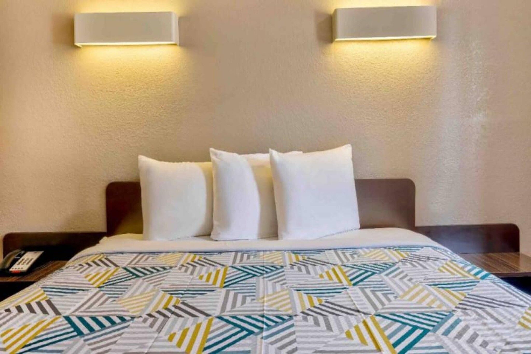 Habitación de hotel con cama, almohadas, colcha estampada y dos lámparas de pared.