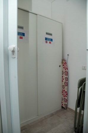 A hallway with a door labeled 'MAGAZZINO' and 'LOCALE TECNICO' signs, a patterned umbrella leaning against the wall, and a chair to the right. - New Listing (Roma)
