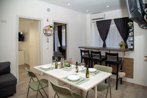 A dining area with a table set for two, chairs, a wall-mounted shelf with decorative items, and an air conditioning unit. - New Listing (Roma)