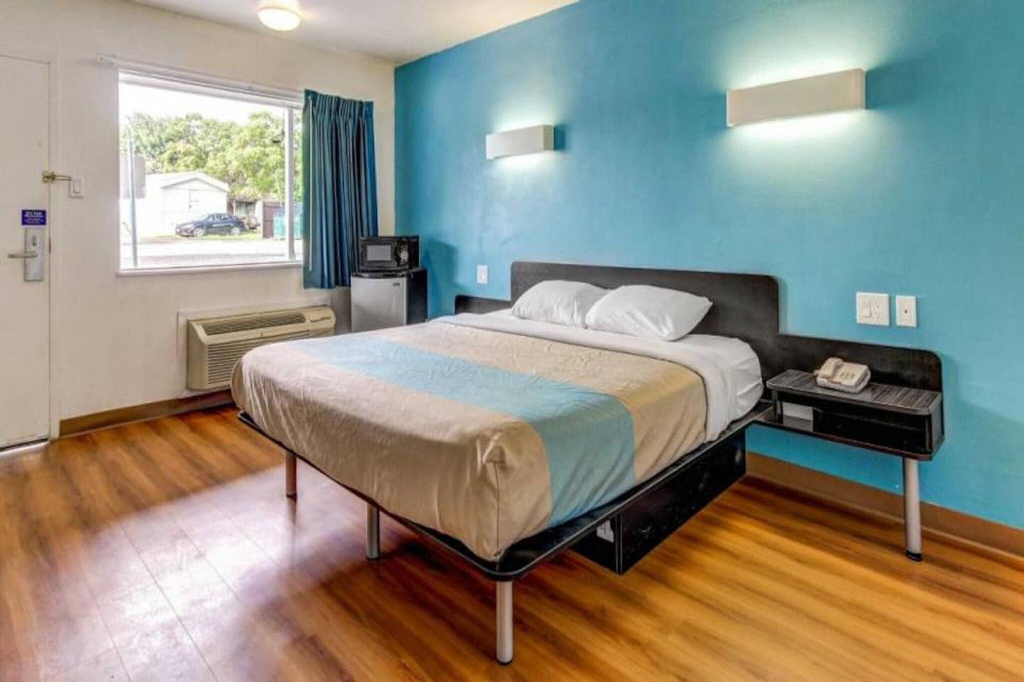 A hotel room with a bed, a nightstand, a heater, a window with curtains, and a blue wall.