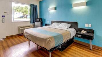 A hotel room with a bed, a nightstand, a heater, a window with curtains, and a blue wall.