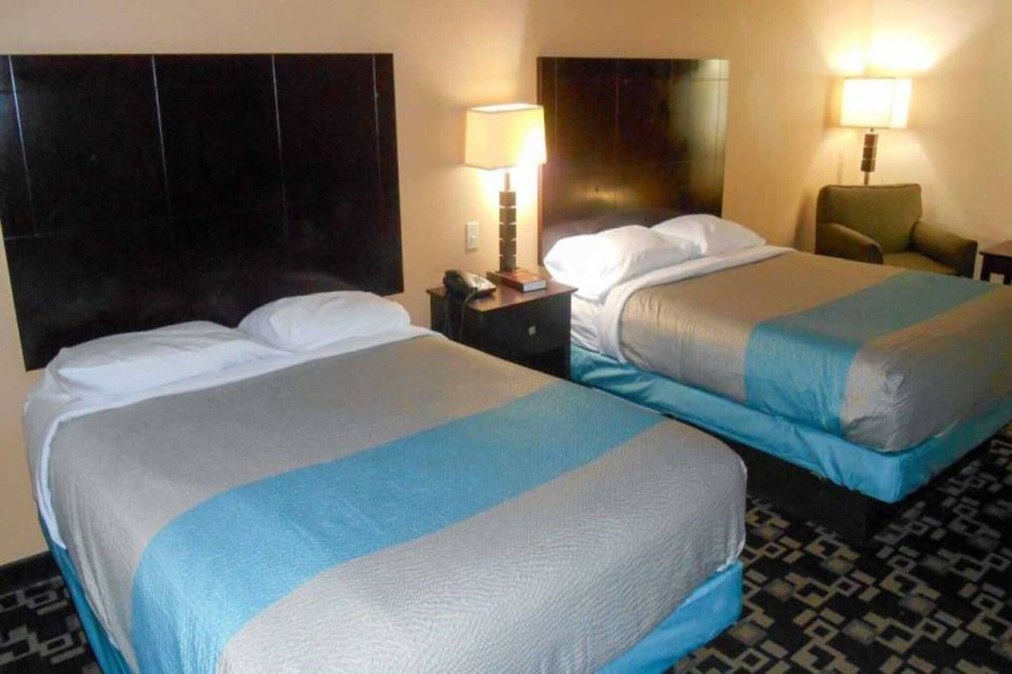Quarto de hotel com duas camas, uma mesa de cabeceira com abajur e uma cadeira.