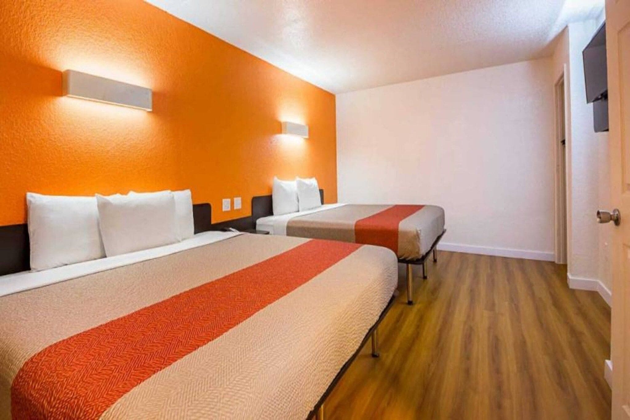 A hotel room with two beds, a wooden floor, and a plain wall.