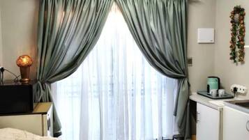 A compact room with a bed, a small table, a window with curtains, a kitchenette, and a wall-mounted light fixture.