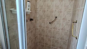 A shower with beige tiles, a glass door, a showerhead, a handheld shower, a towel rack, and a step.