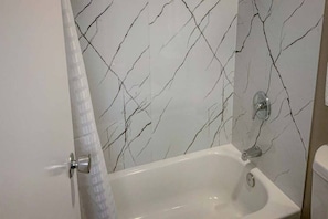 A bathroom with a white bathtub, a shower curtain, and a marble-patterned wall. - 1 King Bed | Non-Smoking, Kitchen (San Angelo)