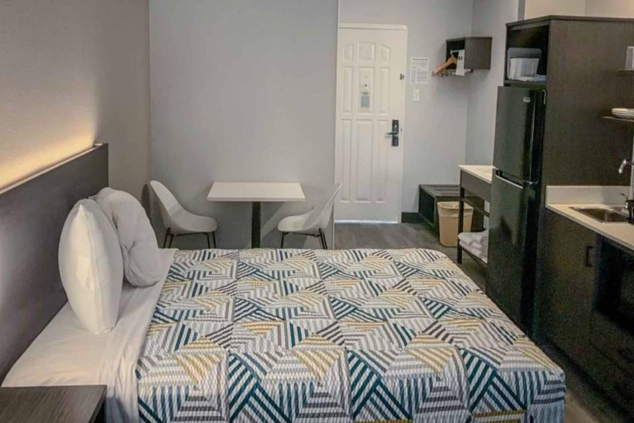 Une chambre d’hôtel avec un lit, deux chaises, une petite table, un réfrigérateur et une porte.