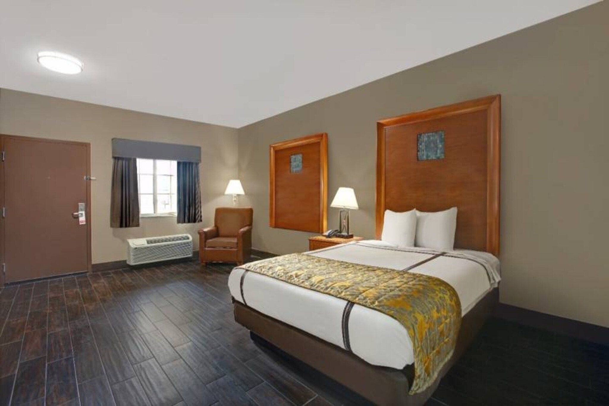 A hotel room with a large bed, a chair, a desk with a lamp, a window with curtains, and a door.