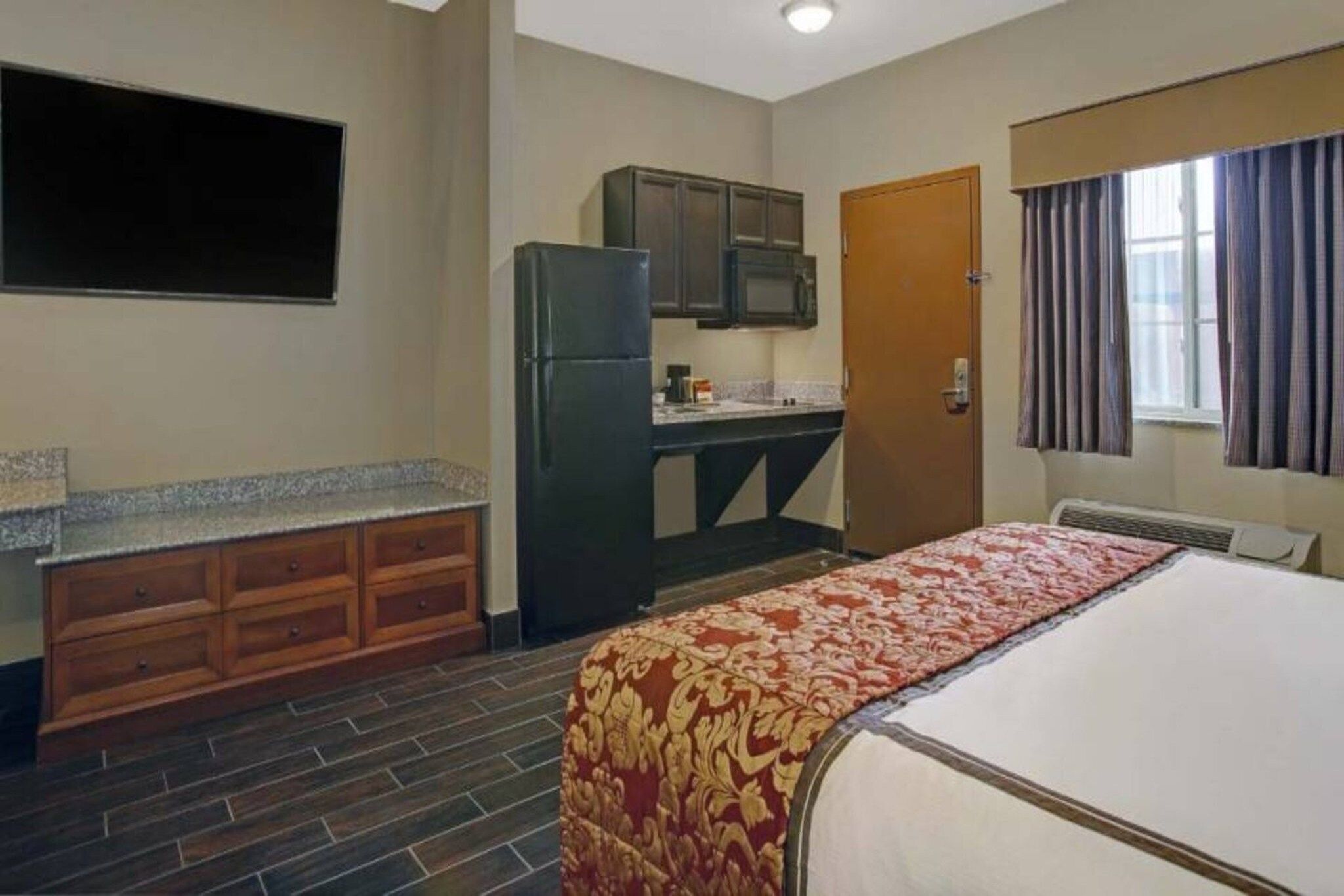 Una habitación de hotel con cama, kitchenette, televisor de pantalla plana y ventana con cortinas.