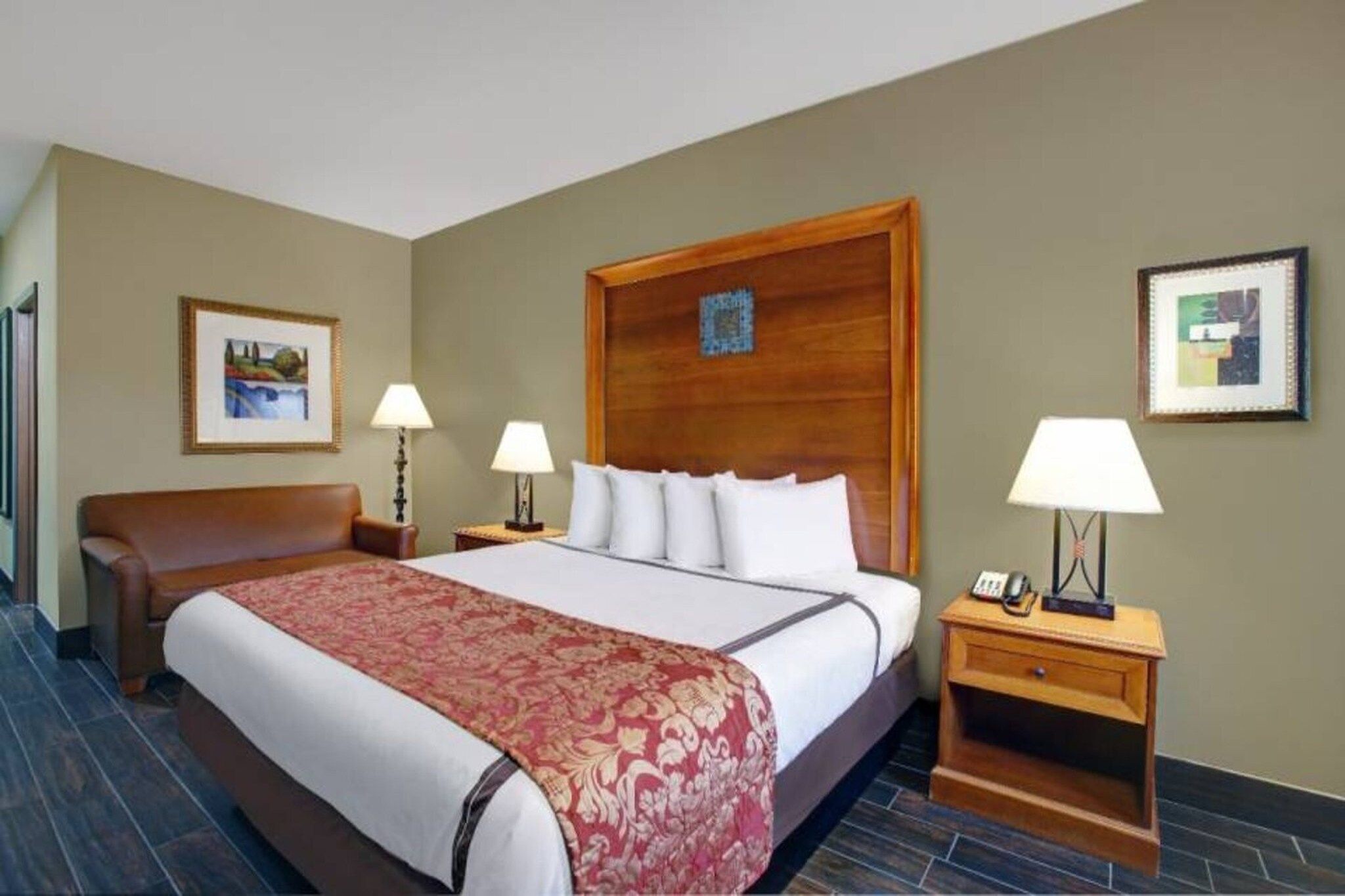 Une chambre d’hôtel avec un grand lit, deux tables de chevet avec des lampes, un fauteuil en cuir marron et deux tableaux encadrés accrochés aux murs.