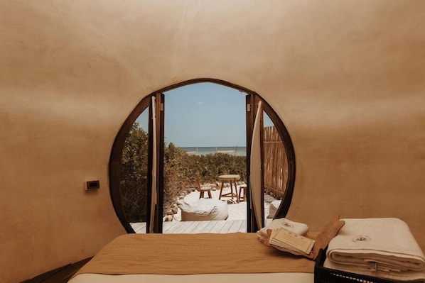 A bedroom with a circular window, a bed, and a view of a beach. - Casa K u Hotel (Dzemul)
