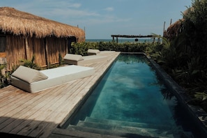 Zona de piscina con terraza de madera, sillas de descanso y una estructura de techo de paja.