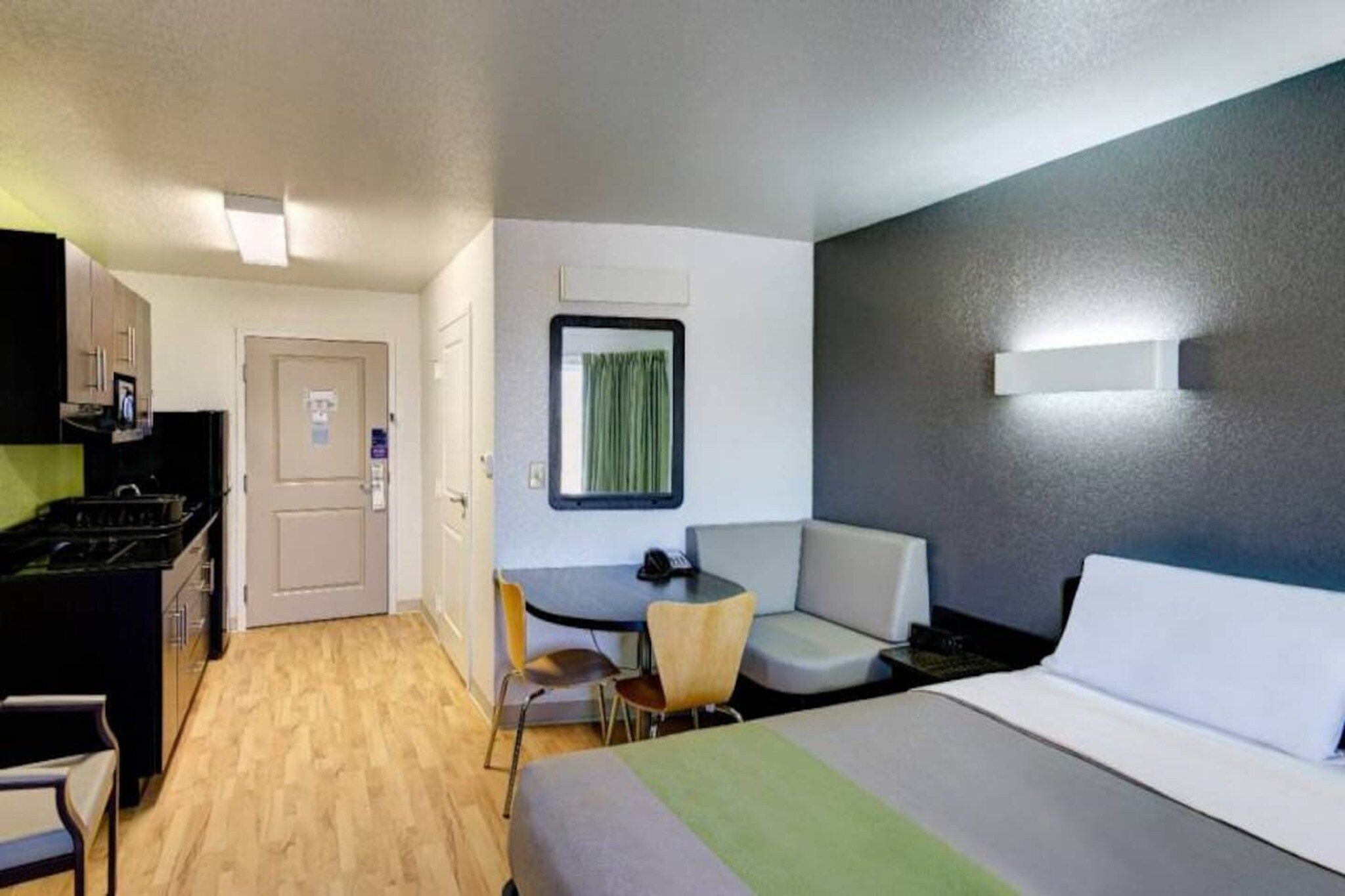 A hotel room with a bed, a desk with a chair, a small kitchenette, and a window with curtains.