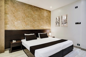 Una habitación de hotel moderna con una cama grande, cabecera de madera oscura, mesita de noche con un teléfono y una lámpara de pared.
