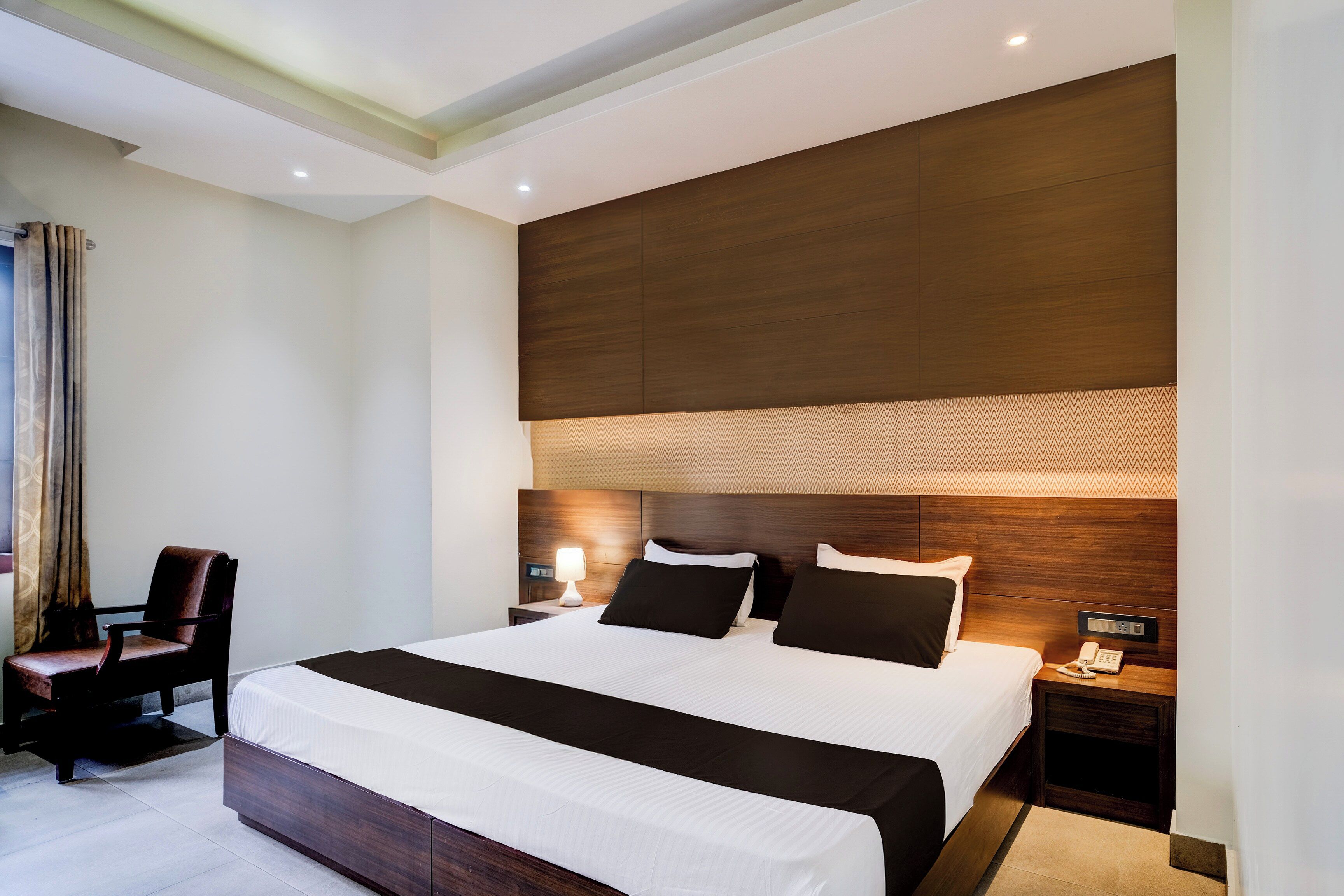 A modern hotel room with a large bed, a bedside table with a lamp, a chair, and a nightstand with a phone.