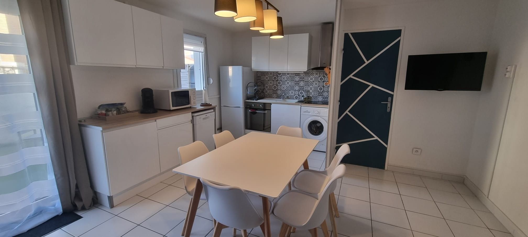 A compact kitchen with a dining table and chairs, a washing machine, and a TV mounted on the wall.