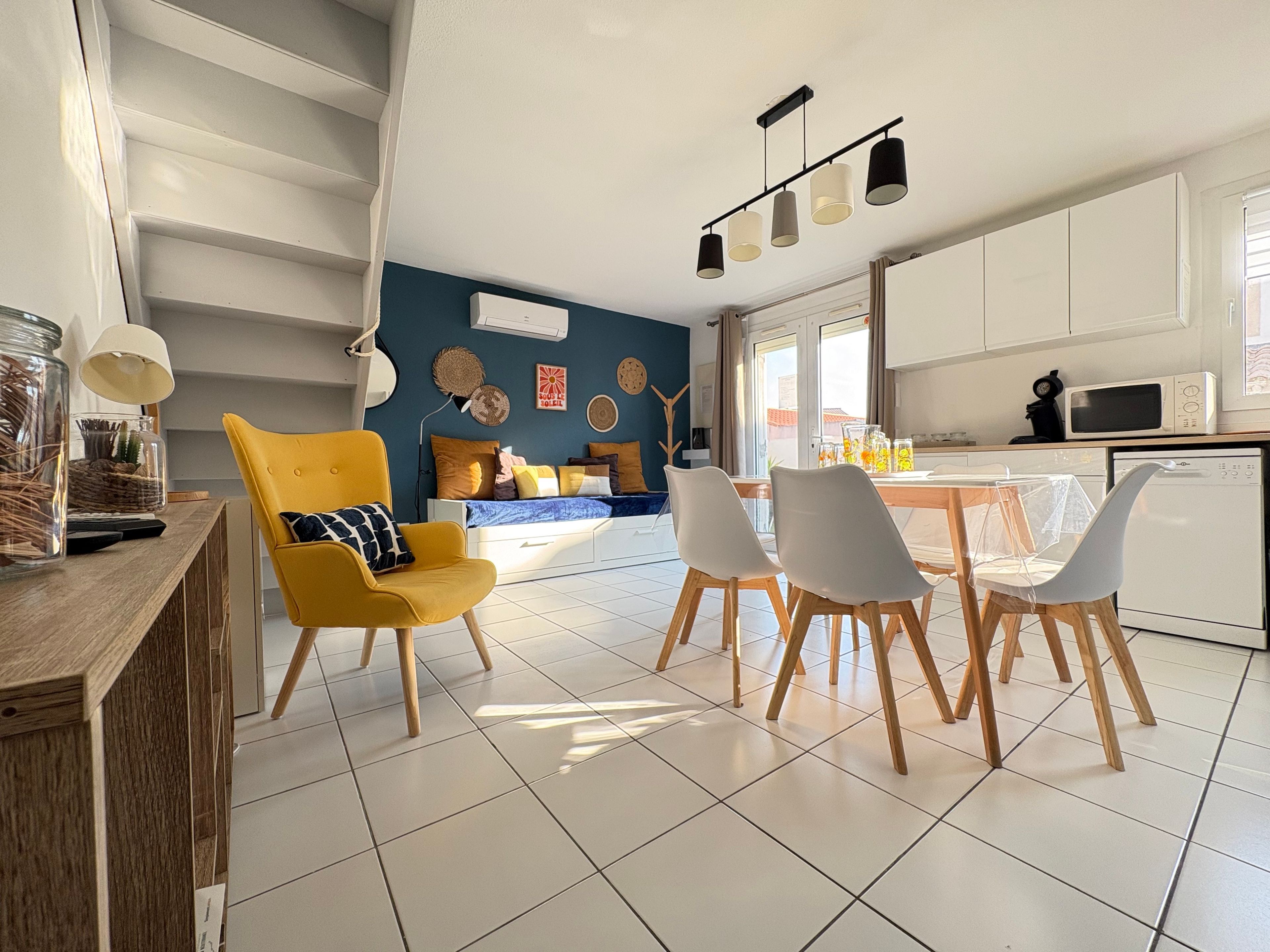 Une cuisine moderne avec des meubles blancs, un coin repas agrémenté d’une chaise jaune et un mur d’accent bleu.