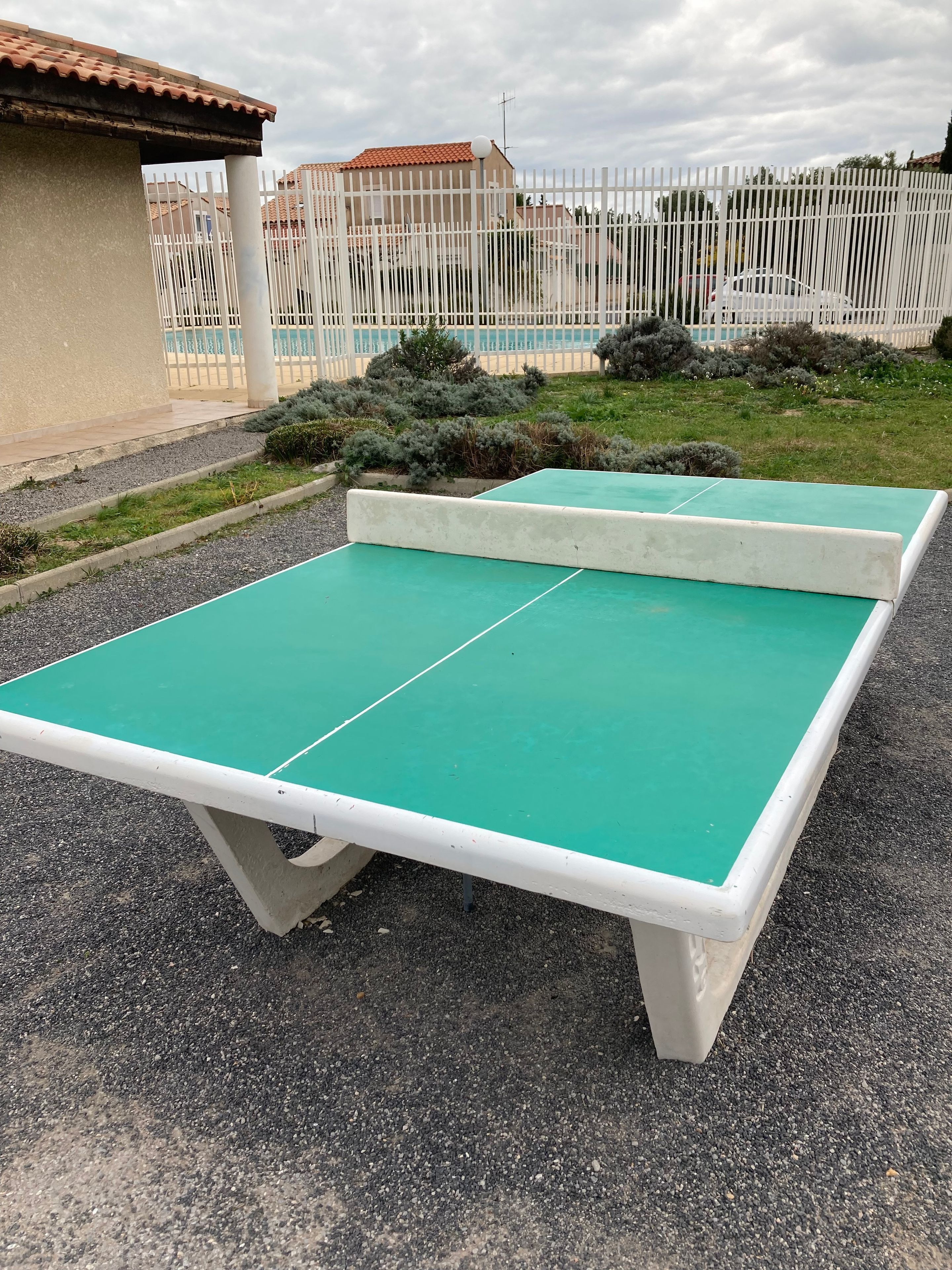 Ein Outdoor-Tischtennis-Tisch mit weißem Rahmen, auf einem Kiesgrund platziert, umgeben von einem Zaun und Grünpflanzen.