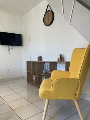 Une pièce avec un fauteuil jaune, une armoire en bois, un téléviseur à écran plat et une horloge murale.