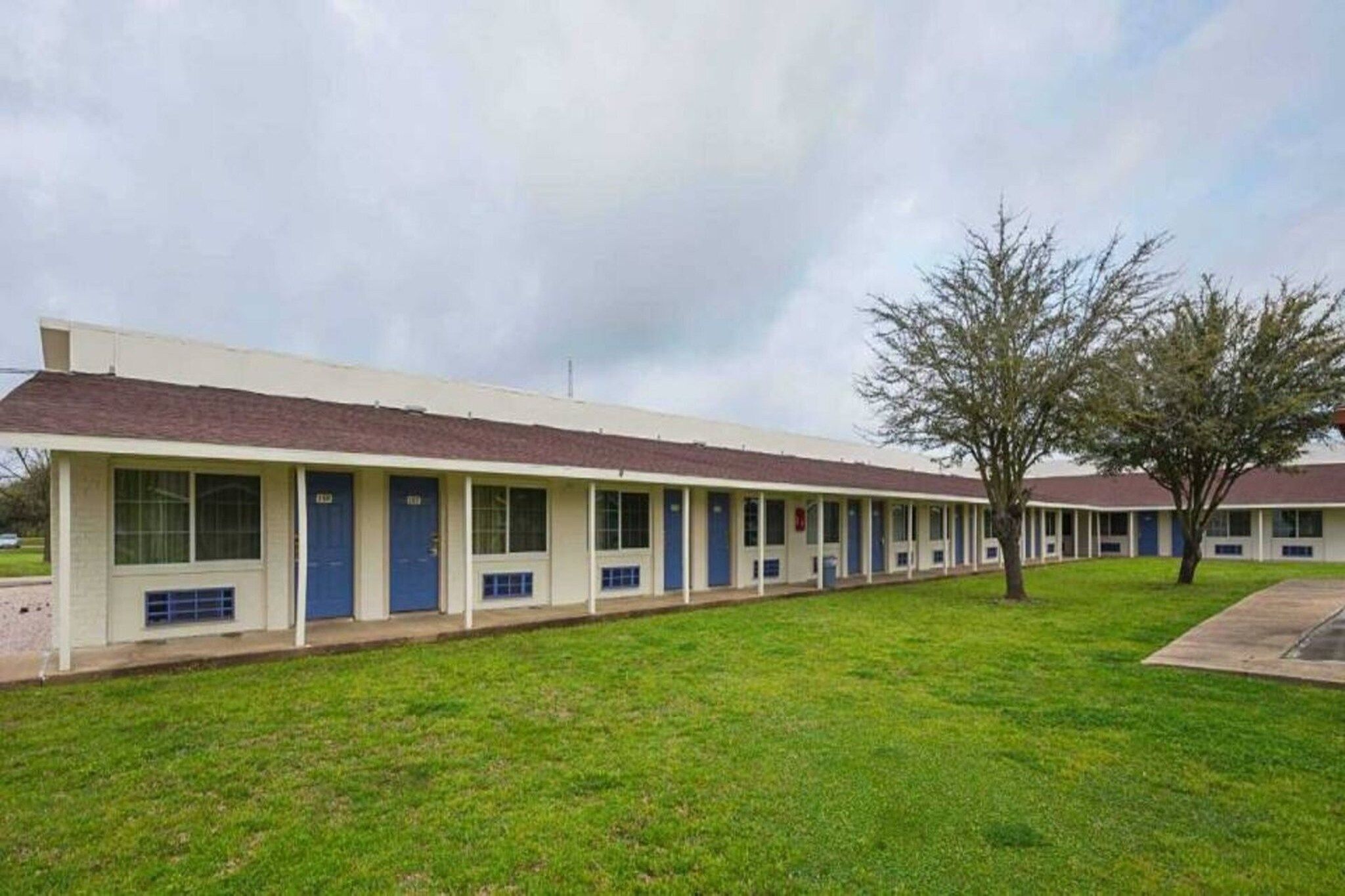 A row of motel buildings with individual rooms, a grassy lawn, and a tree.