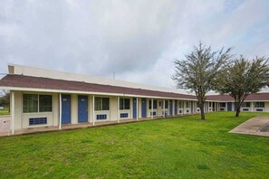 A row of motel buildings with individual rooms, a grassy lawn, and a tree.