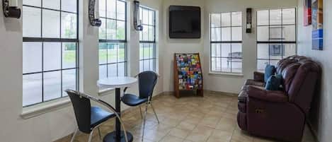 A room with a large window, a flat-screen TV mounted on the wall, a small round table with two chairs, a couch, and a magazine rack.