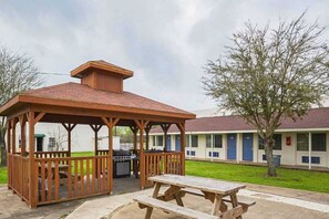 A wooden gazebo with a picnic table in front, a barbecue grill, and a motel building in the background. - 2 Full Beds | Smoking, Micfridge (Madisonville)