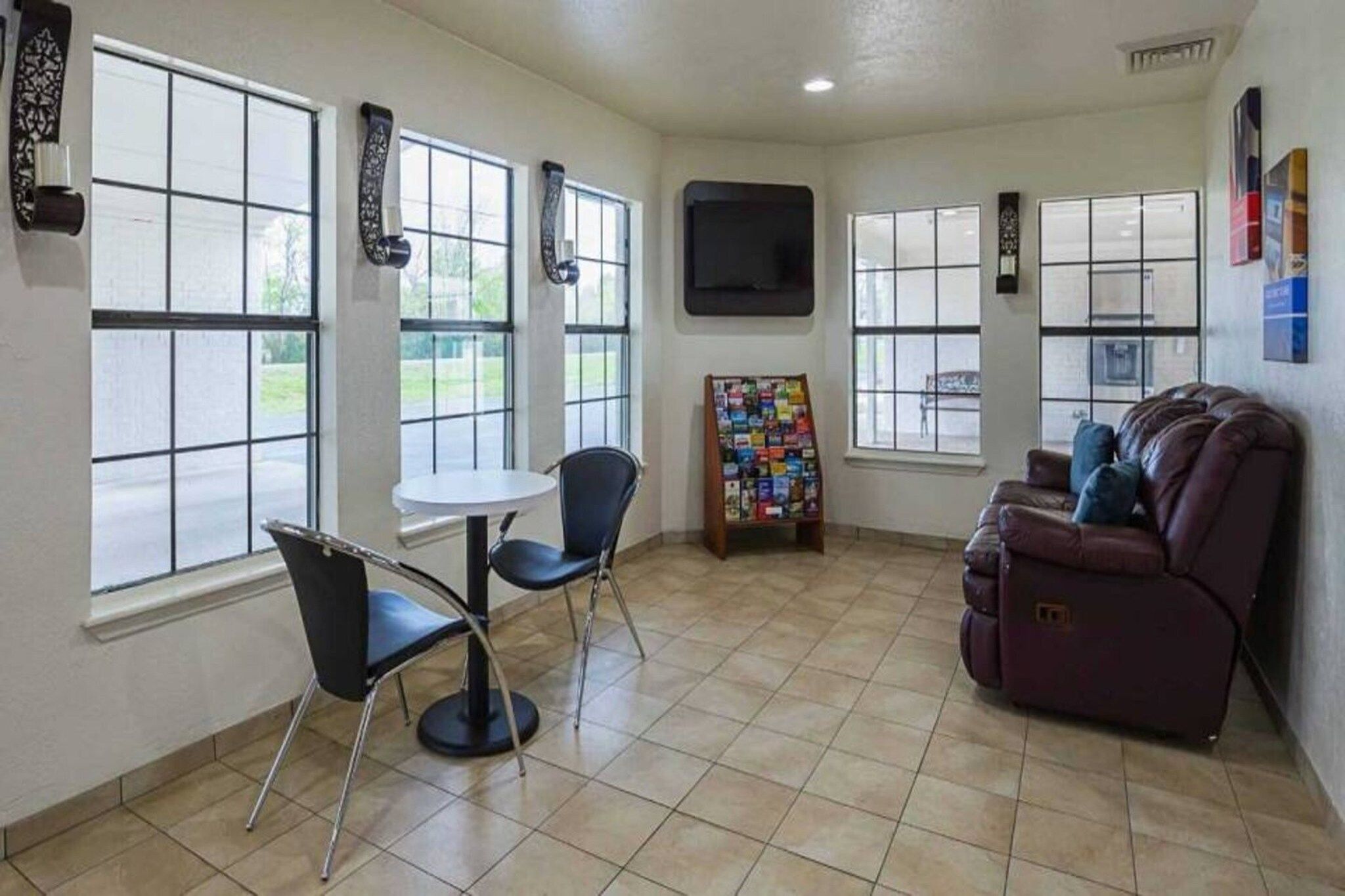 A room with a large window, a flat-screen TV mounted on the wall, a small round table with two chairs, a couch, and a magazine rack.