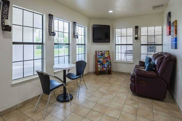 A room with a large window, a flat-screen TV mounted on the wall, a small round table with two chairs, a couch, and a magazine rack.