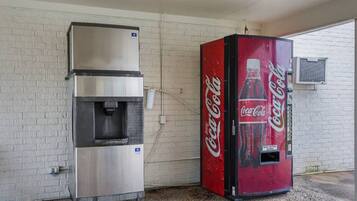 A Coca-Cola vending machine and a stainless steel beverage dispenser against a white brick wall.