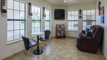 A room with a large window, a flat-screen TV mounted on the wall, a small round table with two chairs, a couch, and a magazine rack.