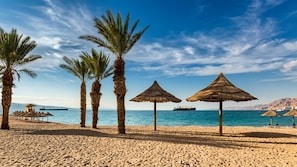 A beach with palm trees, thatched umbrellas, and a calm sea with a ship in the distance. - Seaside suite at 42 Eilat (Eilat)