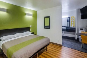 A hotel room with a bed, a small table, a chair, a mirror, and a kitchenette area.