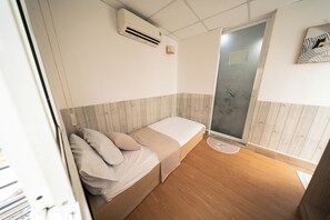 Ein kleines Zimmer mit einem Einzelbett, Holzboden, Wand-Klimaanlage und einer Tür mit Milchglasfenster.