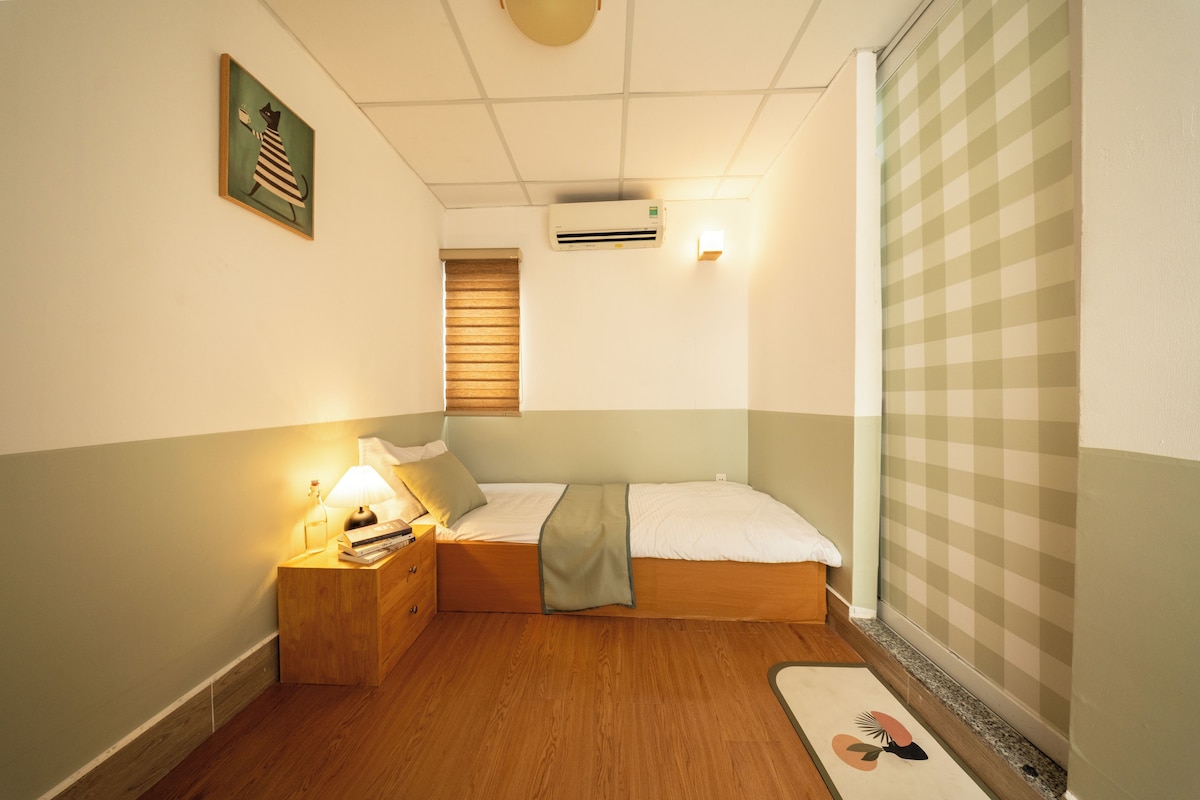 Ein kleines, ordentliches Schlafzimmer mit einem Einzelbett, einem Nachttisch mit Lampe, einer Wand-Klimaanlage und einem Fenster mit Jalousien.