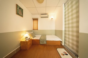 Ein kleines, ordentliches Schlafzimmer mit einem Einzelbett, einem Nachttisch mit Lampe, einer Wand-Klimaanlage und einem Fenster mit Jalousien.