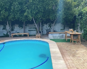 A swimming pool with a tiled edge, surrounded by a brick patio and wooden furniture. - Stellenbosch Holiday Home (Stellenbosch)