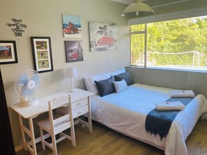 A bedroom with a bed, a desk, a chair, a fan, and a window with a view of trees. - Stellenbosch Holiday Home (Stellenbosch)