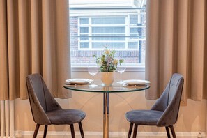 A dining area with two chairs, a glass table, a vase with flowers, and a window with curtains. - Light-filled Studio in Prestigious Chelsea (London)