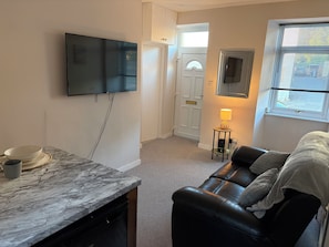 Ett vardagsrum med en soffa, en tv som är monterad på väggen, ett litet bord med en lampa och en dörr som leder till ett annat rum.