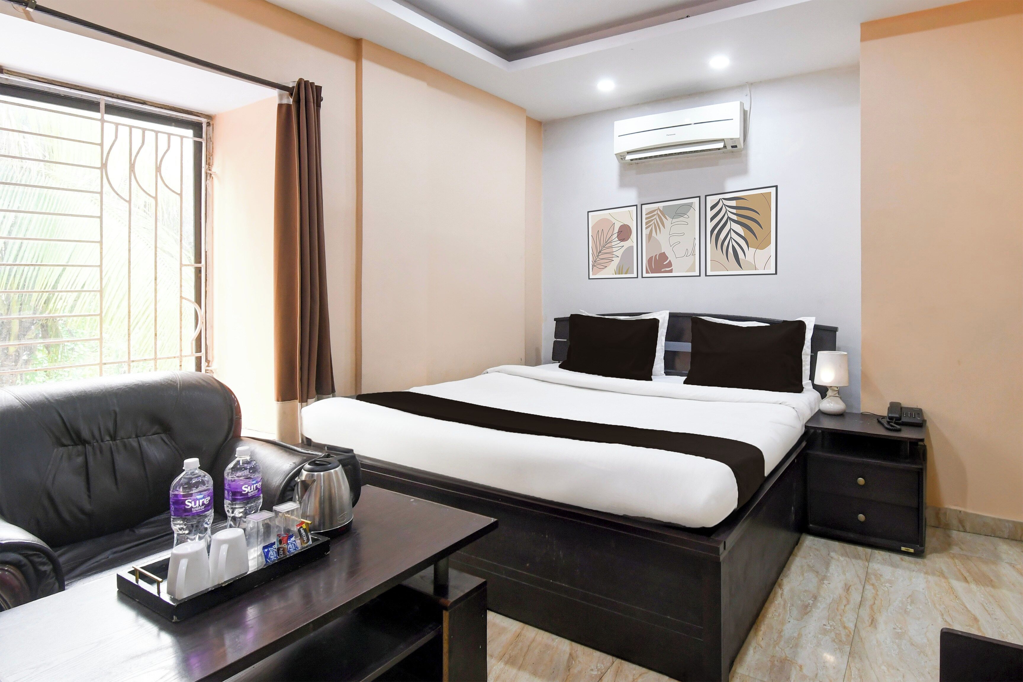 A hotel room with a bed, a desk with a tray of toiletries, a chair, a bedside table with a lamp, and a wall-mounted air conditioner.