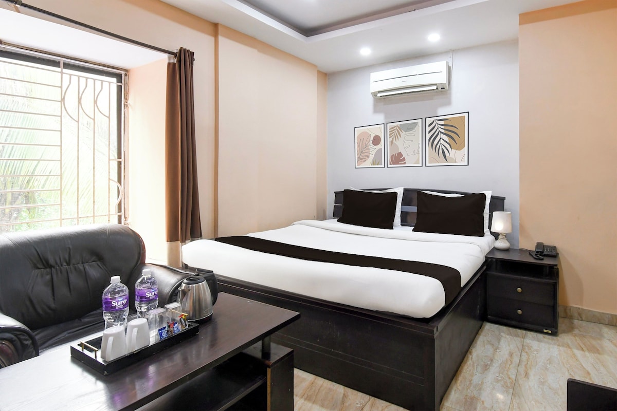A hotel room with a bed, a desk with a tray of toiletries, a chair, a bedside table with a lamp, and a wall-mounted air conditioner.