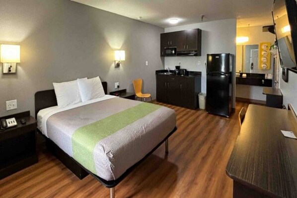 A hotel room with a bed, a desk, a chair, a kitchenette, and a refrigerator.