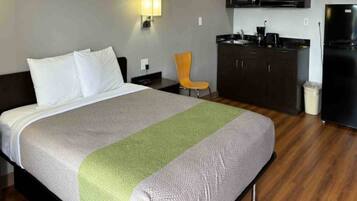 A hotel room with a bed, a chair, a kitchenette, and a trash bin.