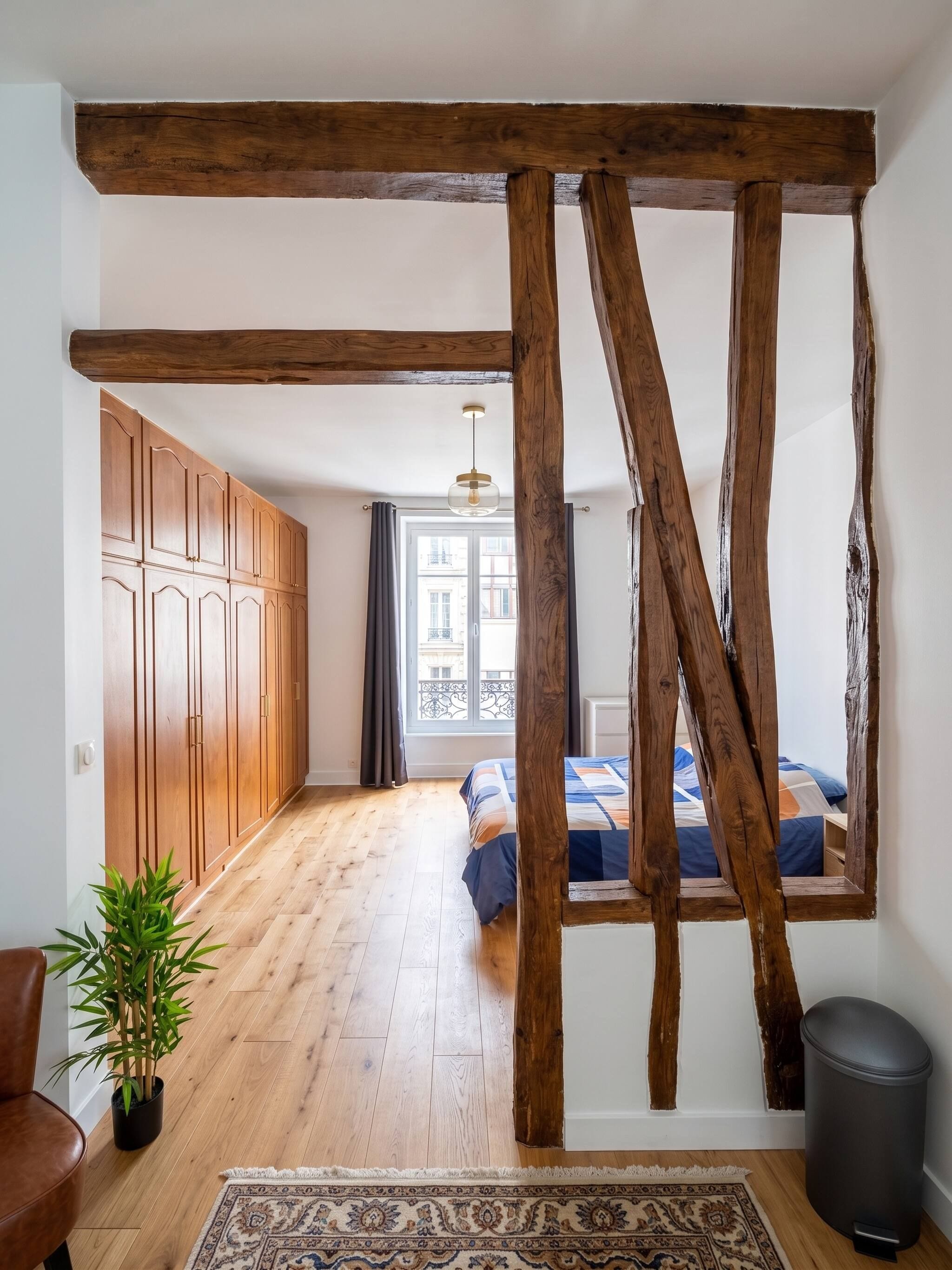 Une pièce avec une poutre en bois au plafond, un lit recouvert d’une courtepointe bleue et orange, une plante en pot, un fauteuil en cuir et un tapis à motifs.