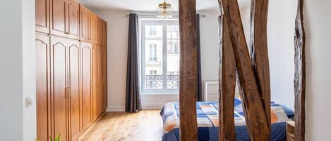 Une pièce avec une poutre en bois au plafond, un lit recouvert d’une courtepointe bleue et orange, une plante en pot, un fauteuil en cuir et un tapis à motifs.