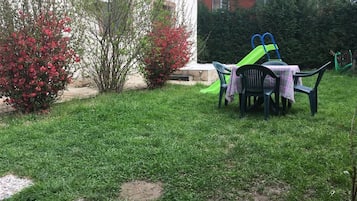 Ein grünes, grasbewachsenes Areal mit einem Tisch, Stühlen, einer grünen Rutsche und roten, blühenden Büschen.